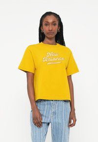 Kollane puuvillane T-särk lühikeste varrukatega ja ümmarguse kaelusega, millel on silmatorkav retrostiilis "New Balance Athletics" logo kreemjas värvuses.