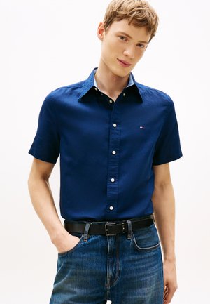 Junger Mann trägt ein marineblaues kurzärmliges Hemd mit Knöpfen und blaue Jeans mit einem schwarzen Gürtel, steht vor einem einfarbigen hellen Hintergrund.