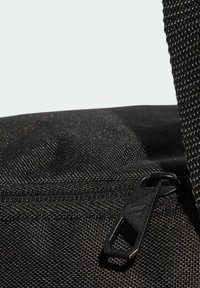 Detalle de una bolsa de tela negra que muestra un material texturizado, un cierre de cremallera y una correa resistente. Cuenta con un tirador de cremallera liso con un diseño grabado.