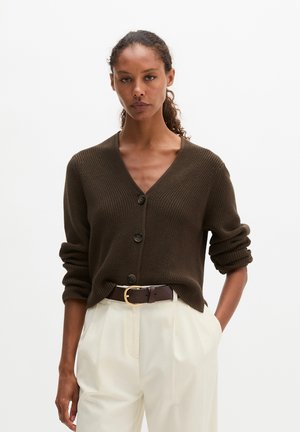 Femme portant un cardigan boutonné marron foncé, un pantalon blanc taille haute et une ceinture marron foncé avec une boucle dorée, posant avec une main dans la poche.