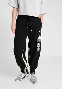 Zwarte joggers met gele zijstrepen, tailleband met trekkoord en meerdere logo's, gecombineerd met grijze sportschoenen. Zachte stof en een relaxte pasvorm.