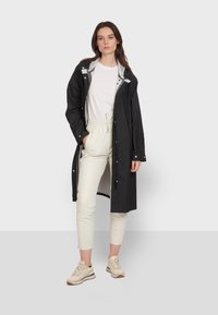 Czarny wodoodporny długi płaszcz z jasnoszarym podszewką, z zatrzaskami i sznurkiem w talii, zestawiony z jasnoszarymi spodniami i trampkami.