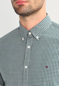 Man in een groen en wit geblokt overhemd met knopen en een klein geborduurd vlaglogo op de borst.