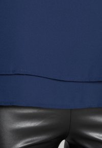 Haut bleu marine avec une texture lisse, doté d'un design superposé et d'un ourlet droit. Associé à un pantalon en cuir noir serré.