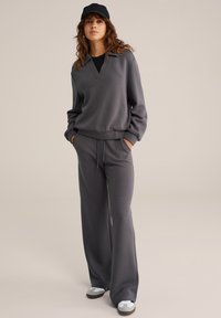 Mujer vistiendo pantalones de pierna ancha gris oscuro y una blusa de manga larga a juego con cuello, gorra negra, zapatillas blancas, de pie con las manos en los bolsillos.