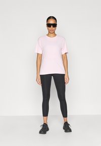 Αθλητική μπλούζα σε ανοιχτό ροζ χρώμα που απορροφά την υγρασία, με κοντά μανίκια, συνδυασμένη με μαύρο εφαρμοστό κολάν και μαύρα αθλητικά παπούτσια.