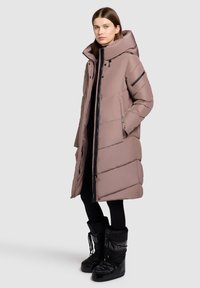 Padded long coat in mauve with a high collar, zip front, side pockets, and diagonal quilted pattern. Worn with black winter boots.