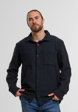 Camicia button-up blu scuro, tessuto texture, due tasche frontali, colletto, maniche lunghe, vestibilità comoda, bottoni visibili.