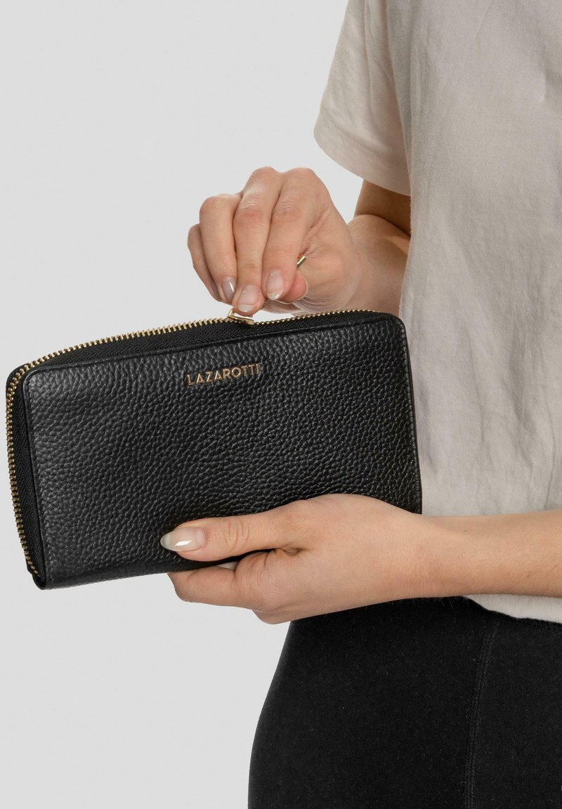 Cartera de cuero negro con un acabado texturizado, cierre de cremallera y marca en tono dorado. Sostenida en una mano, mostrando su diseño delgado y forma.