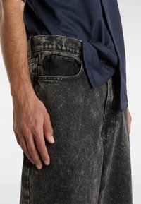 Jeans de mezclilla negra con un aspecto descolorido y desgastado. Presentan un bolsillo frontal para la mano y costuras visibles. Combinados con una camisa texturizada de color azul oscuro.