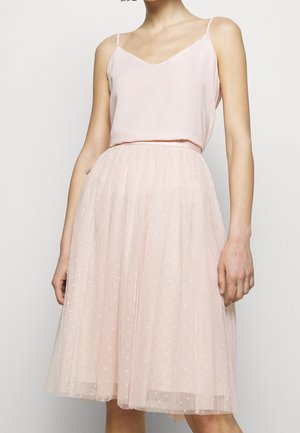Vrouw draagt een lichtroze mouwloos top en een knielange roze tule rok met kleine sterrenpatronen op een eenkleurige achtergrond.