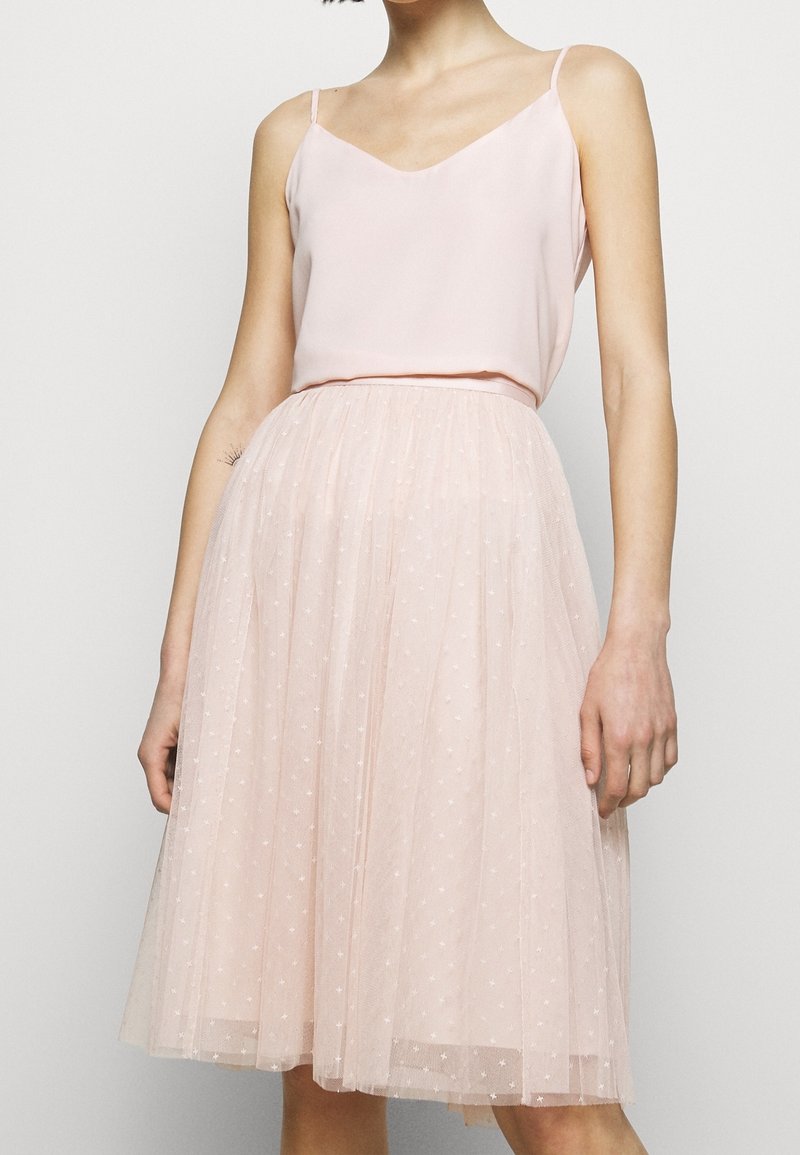Femme portant un haut sans manches rose clair et une jupe en tulle rose longueur genoux avec de petits motifs d'étoiles sur un fond uni.