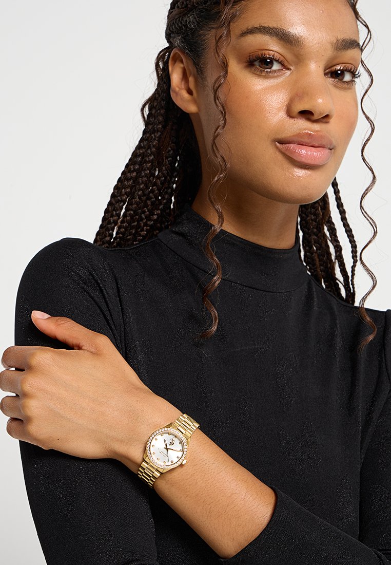 Montre en or avec un cadran rond, un fond blanc et des accents en cristal. Bracelet en métal maillé. Portée sur une main portant un haut à manches longues noir.