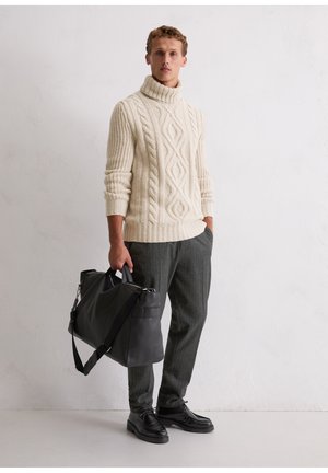 Giovane uomo con maglione crema a trecce con collo alto, pantaloni grigi, scarpe nere, che tiene una grande borsa da viaggio nera in pelle, in piedi contro una parete bianca.