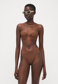 Brun bikinitopp som har en rynket bralette med tynne stropper og matchende høyhåndt bikiniunderdel. Modellen har på seg lyse, retro solbriller.