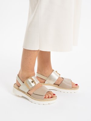 Feet wearing beige and metallic gold open-toe sandals with thick white soles and large gold buckles, paired with a white skirt.