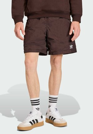 Mand iført sorte Adidas-shorts med hvide Adidas-sneakers og hvide sokker med sorte striber, stående på en lys baggrund.