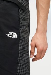 Κοντινό πλάνο του χεριού ενός ατόμου δίπλα σε μαύρο και εμπριμέ παντελόνι με το λογότυπο The North Face στο μηρό.