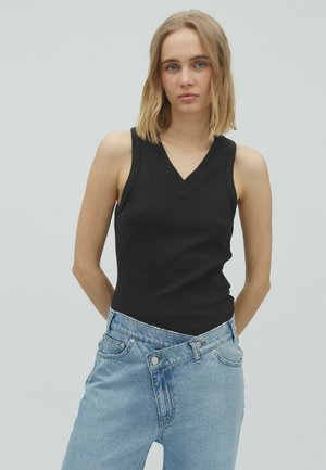 Jeune femme aux cheveux blonds mi-longs portant un top noir sans manches à col en V et un jean bleu asymétrique, debout devant un fond uni.