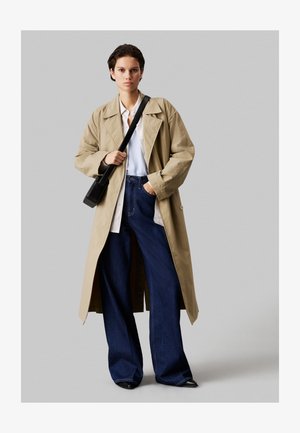 Bež plašč s širokim ovratnikom in gumbnimi detajli, oblečen čez belo srajco in temno modre široke hlače; črna torbica z naramnicami dopolnjuje videz.