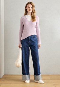 Pulover texturat în nuanța lavandă, cu gât în V, asortat cu blugi evazați de culoare albastră, cu tivuri din denim deschis și adidași albi. Poartă o geantă albă.