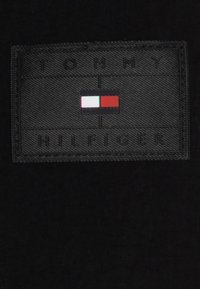 Zwart stoffen label met verhoogde stiksels, met de tekst "TOMMY HILFIGER" en een gekleurd logo van rode, witte en blauwe rechthoeken.