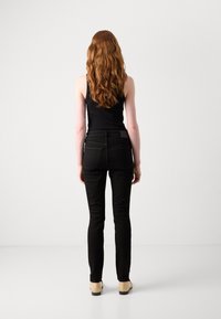 Zwarte slim-fit jeans met een gladde textuur, achterzakken en een subtiele merklabel op de tailleband. Gecombineerd met een zwarte tanktop en beige schoenen.