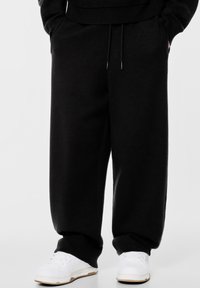 Fekete, laza szabású melegítőnadrág, húzózsin�órral a derékrésznél, bordázott mandzsettákkal és oldalsó zsebekkel, fehér sportcipővel párosítva.