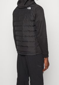 Sort isoleret vest med vandret quiltet mønster, fuld lynlås og logo på venstre skulder. Har et blødt stofhætte og justerbare manchetter.