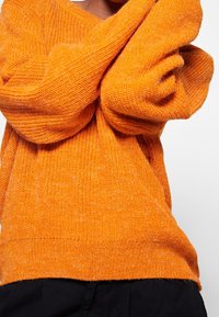 Gestrickter orangefarbener Pullover mit lockerer Passform, strukturiertem Stoff, weiten Ärmeln und V-Ausschnitt. Weiche, flauschige Textur ohne erkennbare Muster.