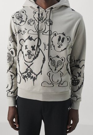 Szara bluza z kapturem z czarnymi kreskówkowymi twarzami i postaciami misia, niektóre z przekreślonymi oczami i oczami w spirali, w komplecie z czarnymi spodniami.