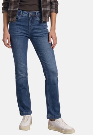 Personne portant un jean bleu droit, des baskets blanches et beiges, et une veste à carreaux avec un haut foncé.