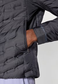 Black puffer jacket featuring a quilted design, zippered pocket, and cuffs with blue "Therma Fit ADV" text. Smooth, lightweight material.