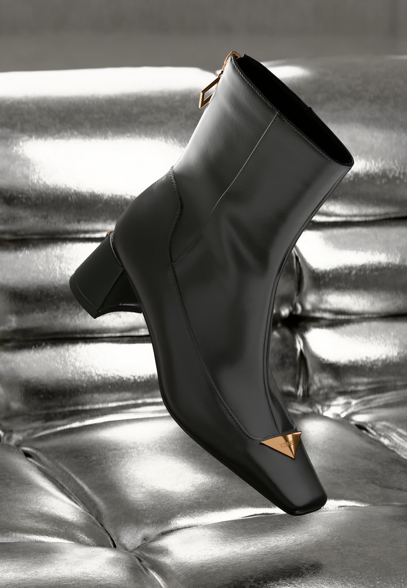 Stivaletto in pelle nera con punta quadrata, accentatura triangolare dorata e tacco medio. Tessuto liscio su uno sfondo argentato lucido.