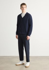 Σκούρο μπλε πλεχτό ζακέτα με υφή, με V-κάτω, και κούμπωμα, συνδυασμένη με σκούρες παντελόνες και λευκά sneakers.