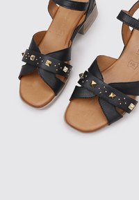 Sandales en cuir noir avec un design de lanières croisées, ornées de clous dorés et d'un talon bloc, avec une semelle intérieure beige et une surface texturée.