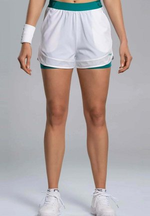 Atleet die witte en groene gelaagde sportshorts, een wit polsbandje en witte sportschoenen draagt, staand tegen een effen grijze achtergrond.