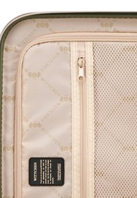 Intérieur d'une valise rigide verte, avec un revêtement en tissu beige à motif de logo, une poche zippée en mesh et une petite étiquette noire.