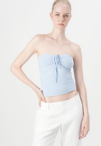 Top azul claro sem alças com um detalhe de laço no centro, feito de um tecido suave. Usado com calças brancas, contra um fundo liso.