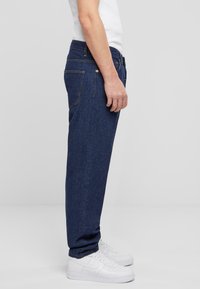 Jeans de mezclilla en azul oscuro, con corte recto y bolsillos estándar. Llevados con zapatillas blancas y una camisa blanca ajustada.
