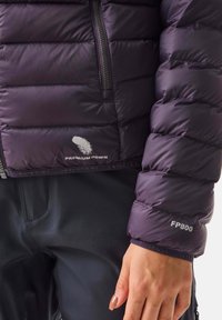 Close-up van een paarse gewatteerde jas met het "Premium Down" logo en "FP800" op de mouw, gedragen met zwarte broek en zichtbare hand.
