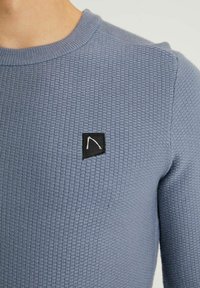 Nära bild av en person som bär en strukturerad ljusblå tröja med en liten svart fyrkantig lapp som har en vit geometrisk design på bröstet.