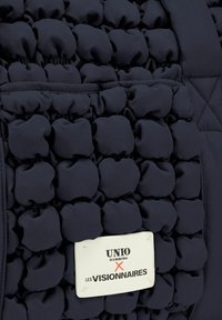Borsa a mano blu navy con un'esterno testurizzato e puffoso. Include un'etichetta con logo rettangolare con testo in bianco e nero ben in evidenza.