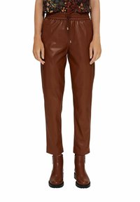 Bruine leren broek met een tapered fit en elastische taille, gestyled met bruine enkellaarsjes. Het materiaal heeft een gladde textuur en een matte afwerking.