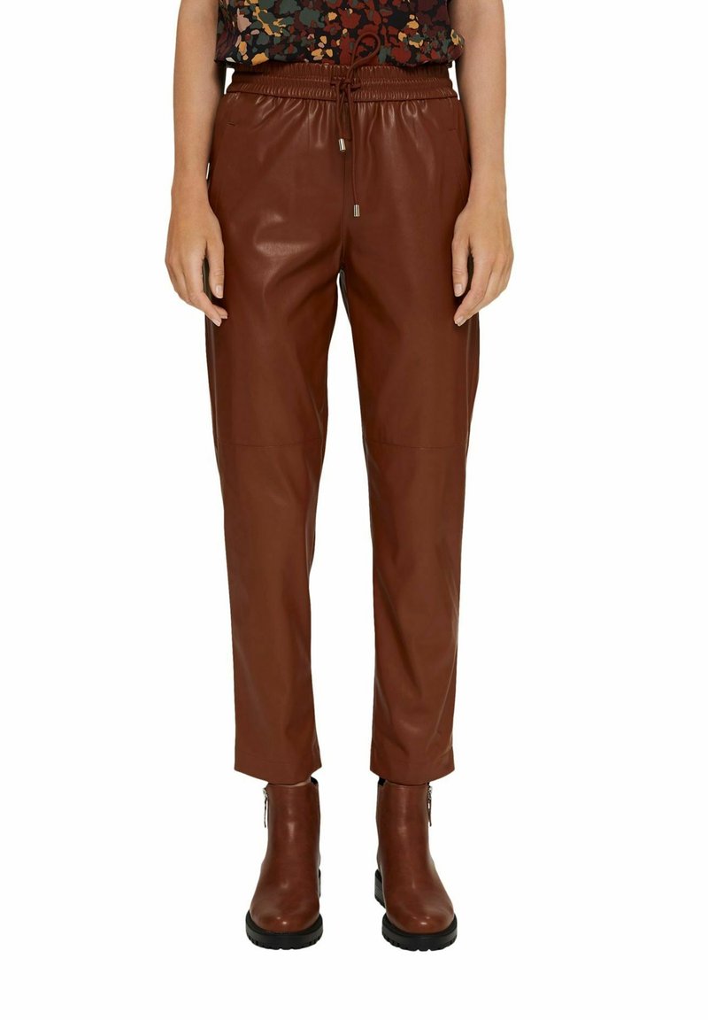 Bruine leren broek met een tapered fit en elastische taille, gestyled met bruine enkellaarsjes. Het materiaal heeft een gladde textuur en een matte afwerking.