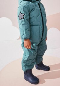 Teal gewatteerde sneeuwpak met een capuchon, voorzien van een grijze sterrenpatch op de arm en marineblauwe rubberen laarzen. Getextureerd, warm en praktisch ontwerp.