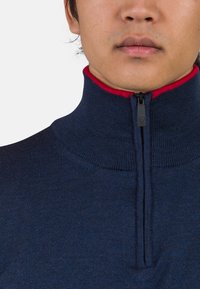 Navy Zip-Pullover mit hohem Kragen und rotem Innenfutter. Hergestellt aus strukturiertem Strickmaterial, mit schwarzem Reißverschluss vorne.