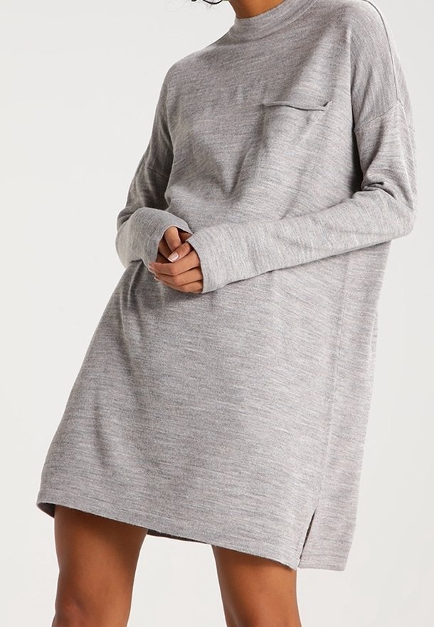 Femme portant une robe tricotée gris clair, ample, à manches longues, avec une petite poche sur la poitrine et une fente latérale, mains jointes à la taille.