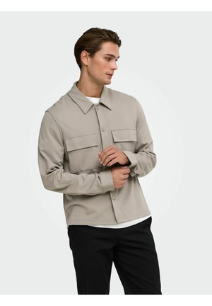 Jeune homme ajustant le poignet d'une veste beige à boutons avec poches poitrine, portant un pantalon noir et un sous-vêtement blanc, regardant sur le côté.