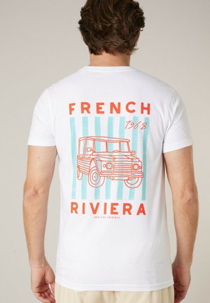Homme portant un t-shirt blanc avec un texte rouge "French Riviera 1968" et un contour rouge d'une voiture vintage sur des rayures verticales bleues au dos.
